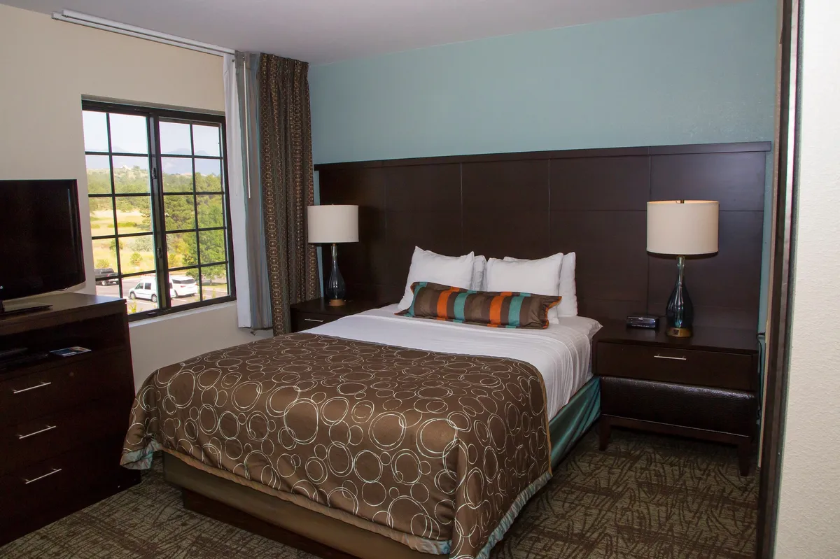 A bright, comfortable bedroom featuring a plush Queen bed with crisp linens, a nightstand with a lamp, a large window allowing natural light, and a television mounted opposite the bed. The calming palette and thoughtful decor create a restful retreat ideal for relaxation.