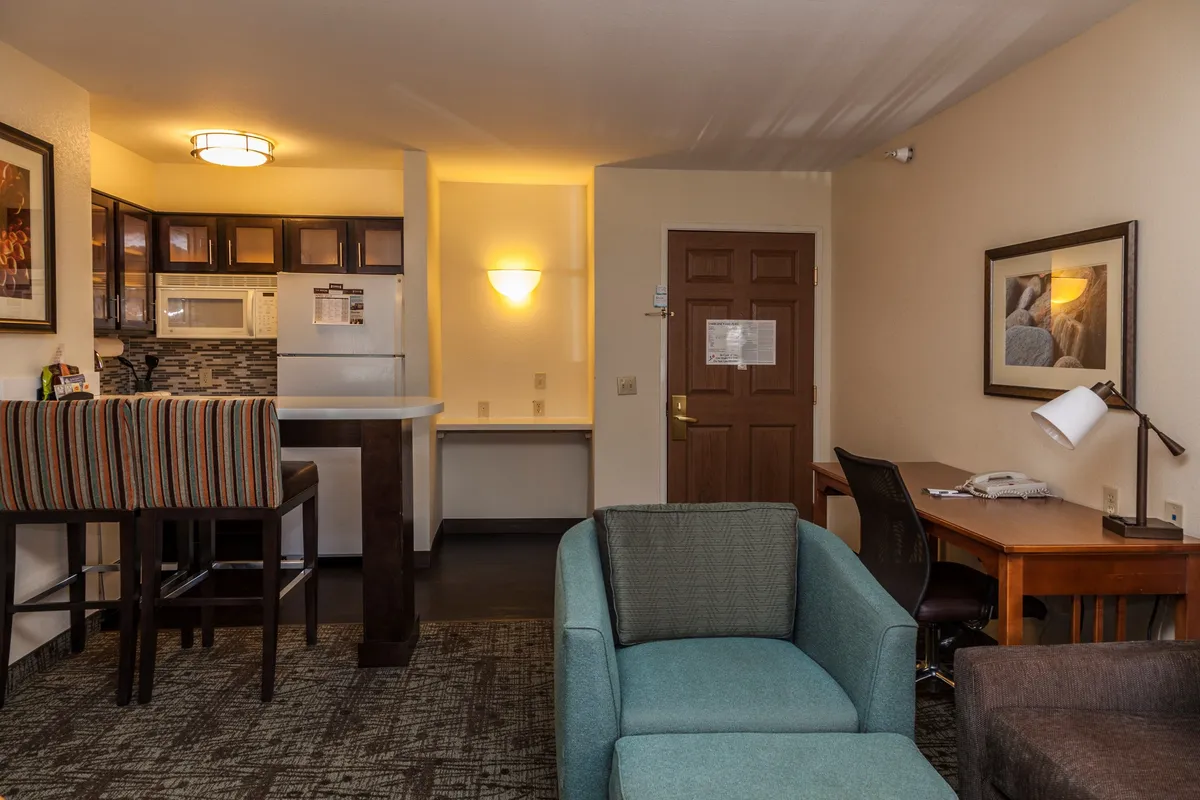 A bright, open living area features a plush sofa facing a flatscreen TV, with a stylish kitchen space visible in the background. Bar stools line the kitchen counter, and a small desk area provides a convenient spot for work or games. The decor is modern and inviting, blending comfort and functionality seamlessly.